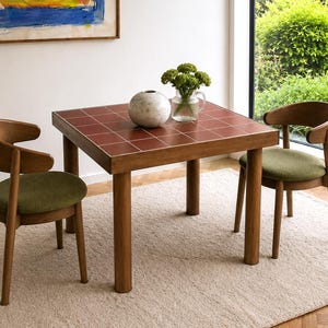 Puede incluir: Una mesa cuadrada con tablero de azulejos rojo oscuro y cuatro patas de madera, acompañada de dos sillas de madera con respaldos curvos y asientos redondos tapizados en verde oliva. Sobre la mesa hay una esfera de mármol blanco y un florero.