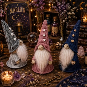Peut inclure: Trois gnomes au crochet en gris, rose poudré et bleu marine. Chaque gnome possède un grand chapeau pointu orné de petits boutons en bois en forme de cœur ou d'étoile, un nez rond et une longue barbe en fausse fourrure blanche. Ils sont debout.