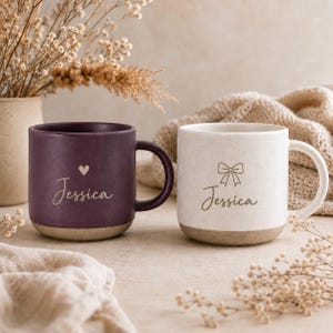 Może przedstawiać: Dwa ceramiczne kubki z matowym wykończeniem, jeden ciemnofioletowy i jeden złamany biały. Fioletowy kubek ma małą ikonę serca nad imieniem Jessica napisanym kursywą. Złamany biały kubek ma ikonę kokardki nad imieniem Jessica napisanym kursywą.