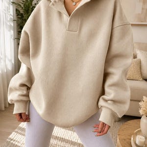May include: An oversized beige hooded sweatshirt featuring a collared neckline with a placket, long sleeves with ribbed cuffs, and a relaxed fit. The garment is paired with white ribbed cycling shorts, creating a neutral-toned ensemble.