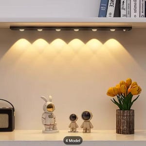 May include: A black linear LED light bar mounted under a shelf, emitting six downward beams of warm yellow light. The light illuminates a surface featuring a rabbit astronaut figurine, two smaller astronaut figurines, and a vase of yellow tulips.