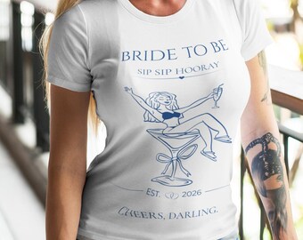 Op de afbeelding: Een wit t-shirt met korte mouwen en blauwe tekst met de woorden BRIDE TO BE, SIP SIP HOORAY, EST. 2026 en CHEERS, DARLING. De afbeelding bevat een lijntekening van een persoon die in een martiniglas zit en een drankje met rietje vasthoudt.