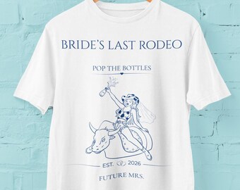 Op de afbeelding: Een wit t-shirt met korte mouwen en een donkerblauwe grafische print. De tekst luidt BRIDE'S LAST RODEO, POP THE BOTTLES, EST. 2026 en FUTURE MRS. De afbeelding toont een persoon met een cowboyhoed en sluier op een mechanische stier.