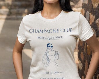 Op de afbeelding: Een crèmekleurig T-shirt met korte mouwen en donkerblauwe tekst: CHAMPAGNE CLUB, BRIDE'S LAST DANCE, EST. 2026 en KISSES FOR THE MRS. De afbeelding bevat een lijntekening van een persoon met een sluier en zonnebril die een glas vasthoudt.