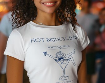 Op de afbeelding: Een wit T-shirt met korte mouwen en de blauwe tekst HOT BRIDE'S CLUB, WIFEY MATERIAL en BRIDE MODE ON. De afbeelding bevat een lijntekening van een persoon in een martiniglas met een drankje, met de tekst EST. 2026 en een hartpictogram.
