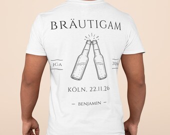Op de afbeelding: Een wit t-shirt met korte mouwen en een zwarte opdruk op de achterkant. Het ontwerp toont twee klinkende bierflesjes met de tekst BRÄUTIGAM erboven, JGA 2026 aan de zijkanten en KÖLN, 22.11.26 met de naam BENJAMIN eronder gedrukt.