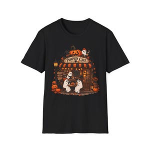 Może przedstawiać: Czarny t-shirt z krótkim rękawem z grafiką kawiarni o nazwie Pumpkin Cafe. Ilustracja przedstawia duchy siedzące przy stolikach, pomarańczowe dynie, markizę w paski i latarnię uliczną, wszystko w ciepłych jesiennych barwach z przodu.