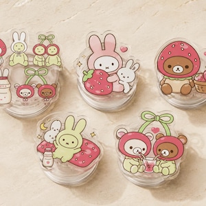 May include: Five clear acrylic phone grips featuring colorful cartoon illustrations of rabbits and bears with strawberries, cherries, and milk bottles. The designs include characters wearing strawberry hats, sharing drinks, and resting on strawberry-patterned cushions.