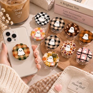 May include: A collection of round, scalloped-edge phone grips featuring a small white ghost character on each. The designs include various patterns such as plaid, floral, and checkered prints in autumn-inspired colors like orange, brown, and black.