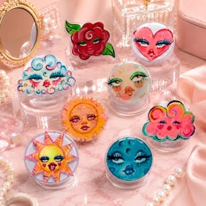 May include: A collection of eight circular phone grips featuring illustrated faces with expressive eyes and lips. Designs include a red rose, a pink heart, a blue cloud, a sun with rays, a moon, and other celestial and floral motifs in vibrant colors.
