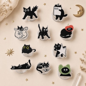 May include: A collection of ten acrylic phone grips featuring various black cat illustrations. The designs include cats in bags, wearing fruit hats, holding stars, listening to music, and playing with boxes, all set on clear circular bases.