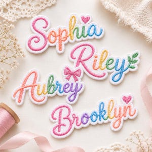 May include: Four embroidered name patches featuring the names Sophia, Riley, Aubrey, and Brooklyn in a colorful gradient script font. Each patch is white with a thick white border and includes decorative elements like hearts and a bow.