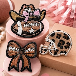 May include: Three embroidered patches featuring a brown cowboy hat with a pink bow atop a football, a brown bow with white football laces, and a football helmet with a brown and black leopard print pattern, all with black stitched borders.