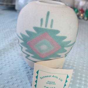 May include: A small, off-white ceramic vase with a rounded shape. It features a turquoise and pink geometric design. The vase has a label that reads "Sunwest Arts, Ltd. Signed Originals NM USA."