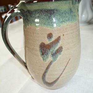 May include: Handmade ceramic pitcher with a large handle. The pitcher has a beige base with a teal and brown glazed top. A teal and brown floral design is painted on the front. The pitcher is a decorative item.