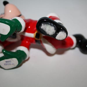May include: A ceramic figurine of a cartoon character in a Santa Claus outfit, lying on its back. The figure is wearing a red suit with white trim, a black belt, green gloves, and black shoes. The figurine is marked "Disney China".