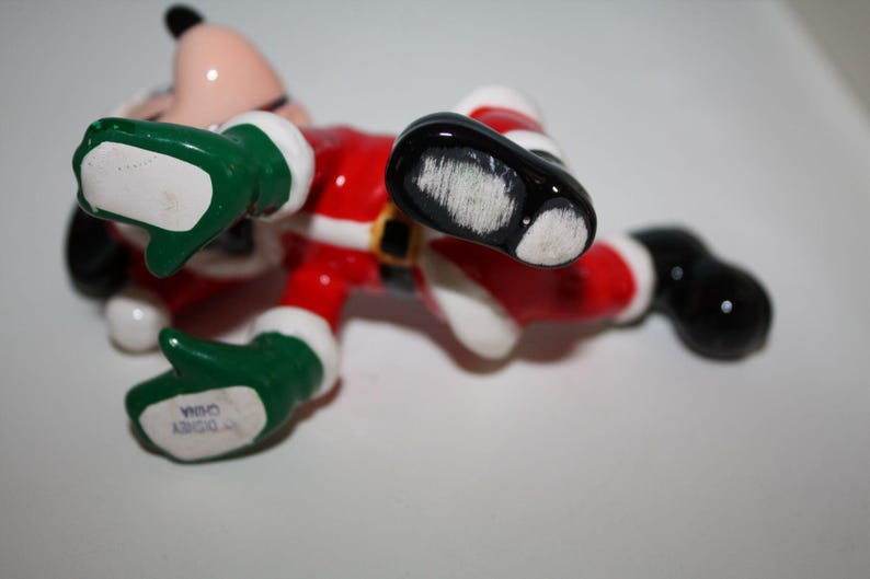 May include: A ceramic figurine of a cartoon character in a Santa Claus outfit, lying on its back. The figure is wearing a red suit with white trim, a black belt, green gloves, and black shoes. The figurine is marked "Disney China".