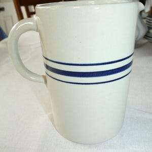 May include: Cream-colored ceramic pitcher with a curved handle and a spout. The pitcher features two horizontal navy blue stripes around the body. A classic, vintage-style kitchenware item.
