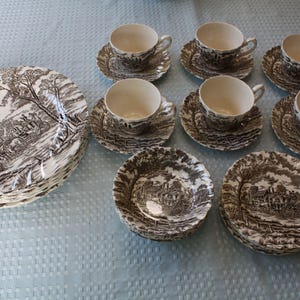 May include: A set of brown and white ceramic dinnerware featuring a detailed pastoral landscape scene. The collection includes a stack of dinner plates, five teacups with matching saucers, and two small bowls, all decorated with a vintage-style print.