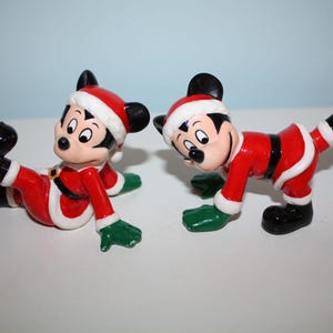 May include: Two Mickey Mouse figurines dressed in Santa Claus outfits. Each figure is wearing a red suit with white trim, a black belt, black shoes, and a Santa hat. The figurines have green gloves and are in playful poses.