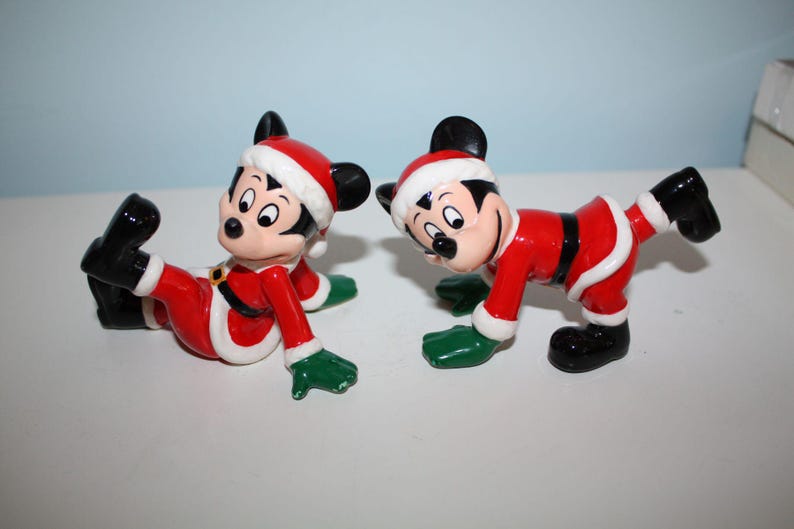May include: Two Mickey Mouse figurines dressed in Santa Claus outfits. Each figure is wearing a red suit with white trim, a black belt, black shoes, and a Santa hat. The figurines have green gloves and are in playful poses.