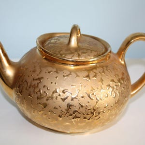 May include: A metallic gold teapot featuring a textured, raised pattern across its rounded body and lid. The teapot includes a curved spout, a matching handle, and a small knob on the center of the lid. The finish has a reflective, shiny quality.