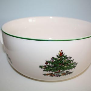 May include: A white ceramic serving bowl with two side handles and a thin green rim. The exterior features a colorful illustration of a decorated Christmas tree with ornaments, a star topper, and wrapped gifts at the base of the tree.