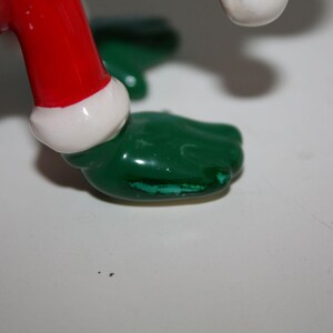 May include: Close-up of a ceramic figurine's foot. The foot is green with a small area of paint loss. The figurine's red leg is visible, trimmed with white. The figurine is on a white surface.