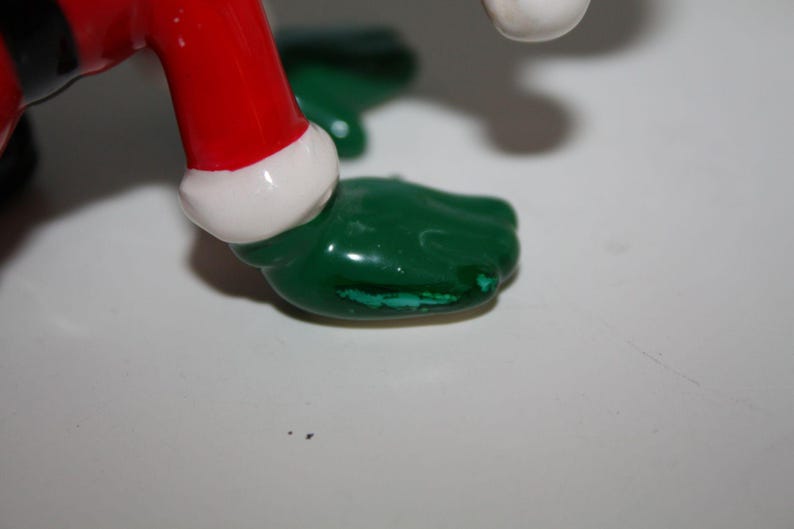 May include: Close-up of a ceramic figurine's foot. The foot is green with a small area of paint loss. The figurine's red leg is visible, trimmed with white. The figurine is on a white surface.