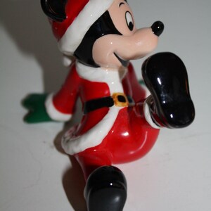 May include: A ceramic Mickey Mouse figurine dressed in a Santa Claus outfit. The figure is predominantly red with white trim, a black belt with a gold buckle, and black shoes. The figure has a cheerful expression.