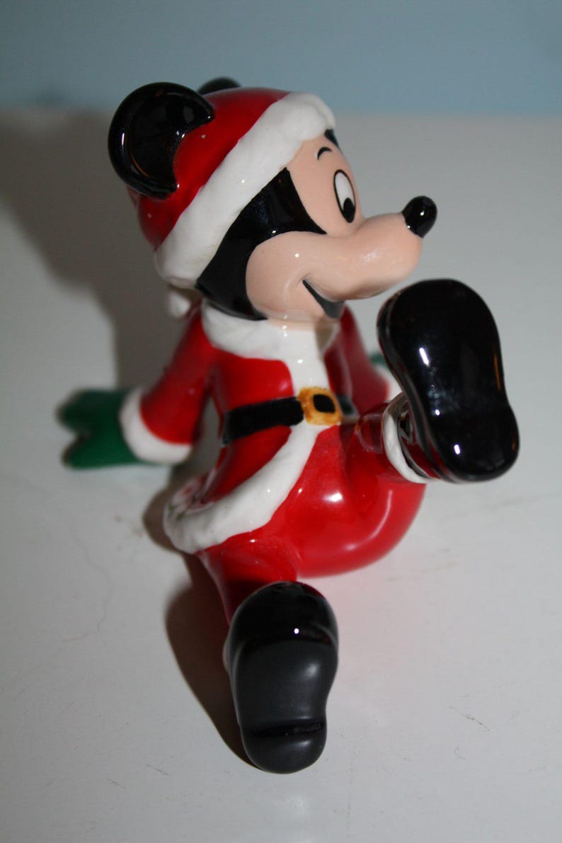 May include: A ceramic Mickey Mouse figurine dressed in a Santa Claus outfit. The figure is predominantly red with white trim, a black belt with a gold buckle, and black shoes. The figure has a cheerful expression.