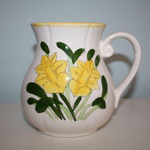 May include: White ceramic pitcher with a yellow rim and handle. The pitcher is decorated with two large yellow daffodil flowers and green leaves. The pitcher is a decorative item.
