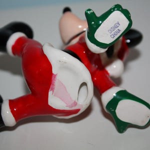 May include: A ceramic Mickey Mouse figurine in a Santa Claus outfit, lying on its back. The figure is red, black, white, and green, with the words "Disney China" on the underside of one hand.