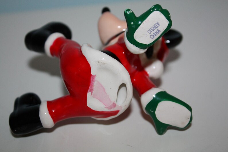 May include: A ceramic Mickey Mouse figurine in a Santa Claus outfit, lying on its back. The figure is red, black, white, and green, with the words "Disney China" on the underside of one hand.