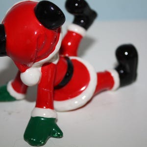 May include: A ceramic Santa Claus figurine in a playful pose. The figure is primarily red with white trim, black boots and gloves, and green mittens. The Santa is lying on its stomach with its legs and arms in the air.