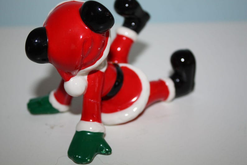 May include: A ceramic Santa Claus figurine in a playful pose. The figure is primarily red with white trim, black boots and gloves, and green mittens. The Santa is lying on its stomach with its legs and arms in the air.
