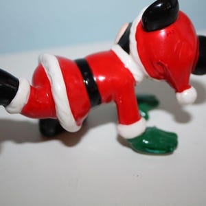 May include: A ceramic figurine of a cartoon character dressed as Santa Claus. The figure is red with white trim, a black belt, and black boots. The character has black ears and green gloves. A festive holiday decoration.