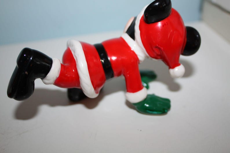 May include: A ceramic figurine of a cartoon character dressed as Santa Claus. The figure is red with white trim, a black belt, and black boots. The character has black ears and green gloves. A festive holiday decoration.