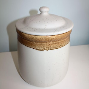 May include: A cylindrical ceramic canister with a speckled off-white finish and a matching lid. The upper portion of the canister features a wide, textured band of mottled brown glaze that creates a distinct contrast against the lighter body.