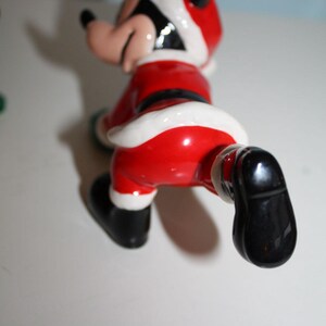 May include: A ceramic figurine of a cartoon character dressed in a Santa Claus outfit. The figure is primarily red with white trim, a black belt, and black shoes. The character's face is visible, with a black hat and ears.