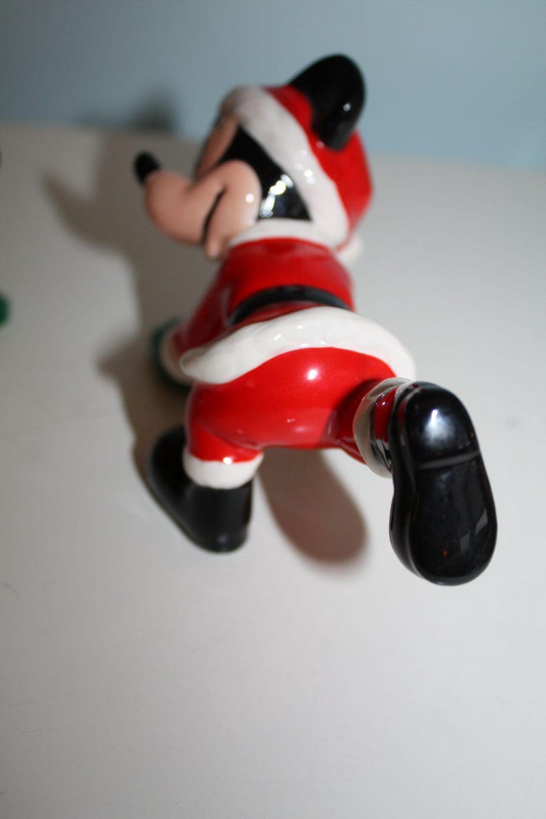 May include: A ceramic figurine of a cartoon character dressed in a Santa Claus outfit. The figure is primarily red with white trim, a black belt, and black shoes. The character's face is visible, with a black hat and ears.