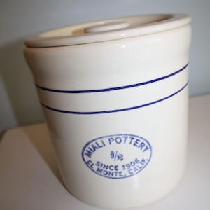 May include: A cream-colored ceramic crock with a matching lid, featuring two horizontal blue stripes around the upper body and a blue stamp that reads MIALI POTTERY 1/2 SINCE 1906 EL MONTE, CALIF. The cylindrical container has a smooth finish.
