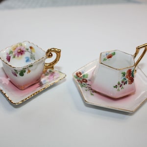May include: Two miniature porcelain teacups and saucers. The cups and saucers are white with a pink ombre effect and gold trim. Floral designs in pink, yellow, and blue decorate the cups and saucers. The handles are gold.