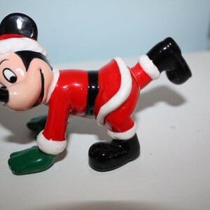 May include: A ceramic figurine of Mickey Mouse in a Santa Claus outfit. The figure is red with white trim, black shoes, and green gloves. Mickey's face is visible, with a black nose and ears, and a red and white Santa hat.
