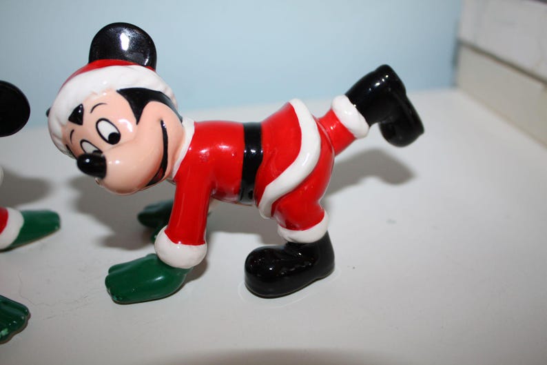 May include: A ceramic figurine of Mickey Mouse in a Santa Claus outfit. The figure is red with white trim, black shoes, and green gloves. Mickey's face is visible, with a black nose and ears, and a red and white Santa hat.