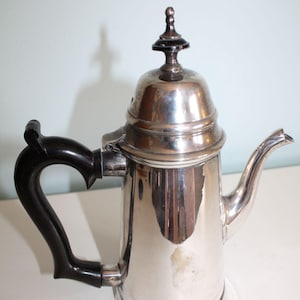 May include: A silver-colored coffee pot with a black handle and lid finial. The pot has a curved spout and a hinged lid. The handle is made of a dark, possibly ebony, material. The pot is likely made of stainless steel.