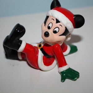 May include: A ceramic figurine of Mickey Mouse dressed in a Santa Claus outfit. The figure is primarily red with white trim, black shoes, and green gloves. Mickey is wearing a Santa hat and has a gold belt buckle.
