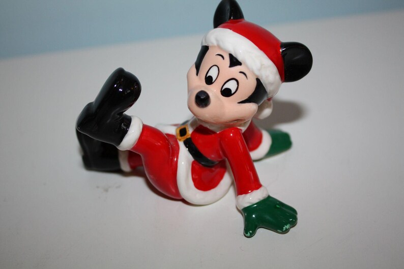 May include: A ceramic figurine of Mickey Mouse dressed in a Santa Claus outfit. The figure is primarily red with white trim, black shoes, and green gloves. Mickey is wearing a Santa hat and has a gold belt buckle.