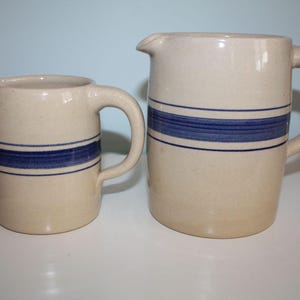 May include: Two ceramic pitchers with a cream-colored base and a blue stripe design. The pitchers have a handle and a spout for pouring. The larger pitcher is approximately 8 inches tall, and the smaller pitcher is about 5 inches tall.