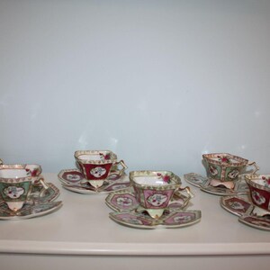 May include: Six vintage teacups and saucers. The teacups are square-shaped with gold handles and a floral design. The saucers are scalloped with matching floral patterns. The teacups have a red, pink, and green color scheme.